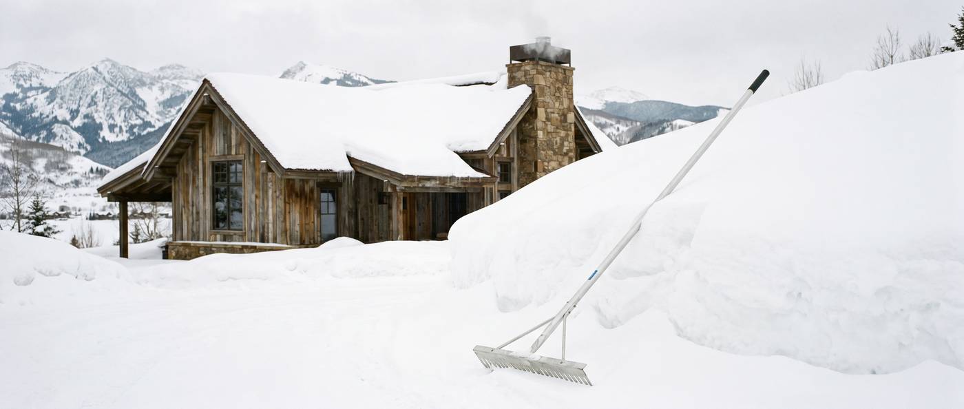 A long-handled aluminum roof rake resting against a snowbank beside a mountain home