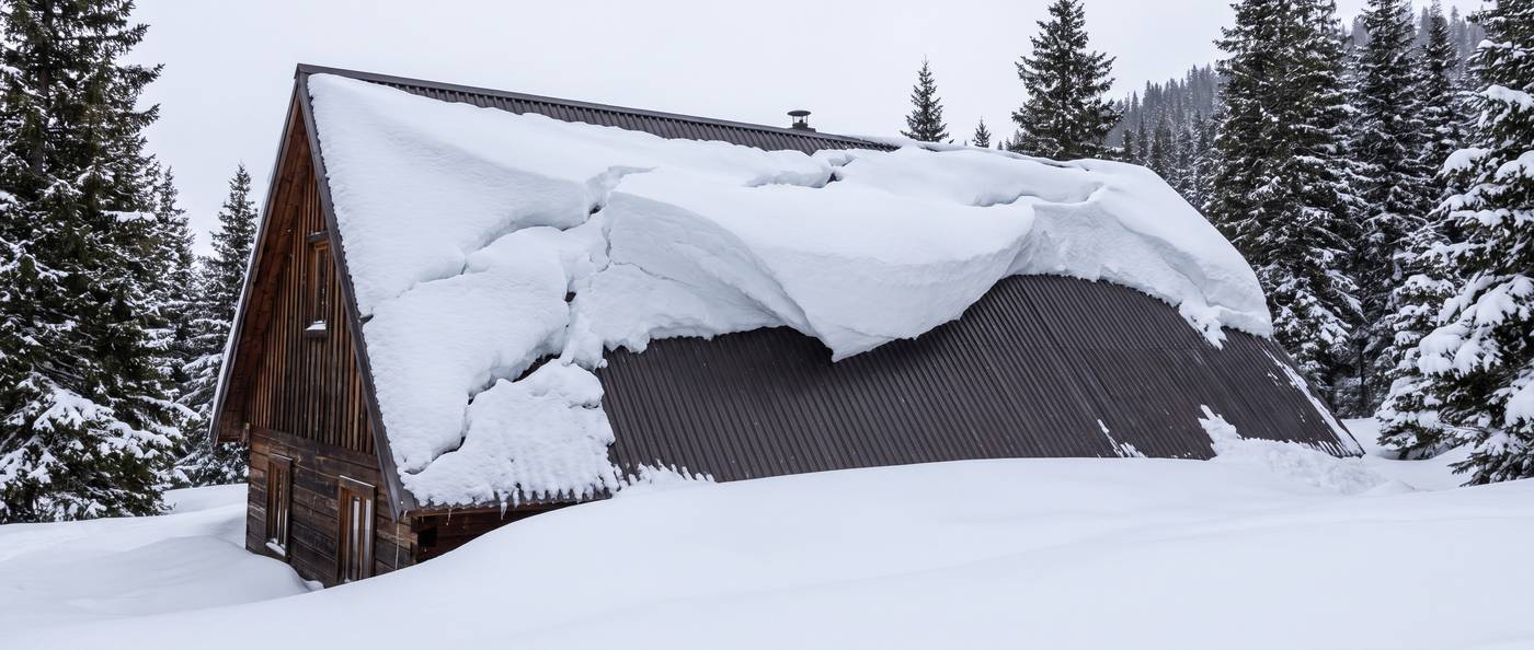 A large cornice of settled snow overhanging the eave of a steep metal roof, cracked and ready to release