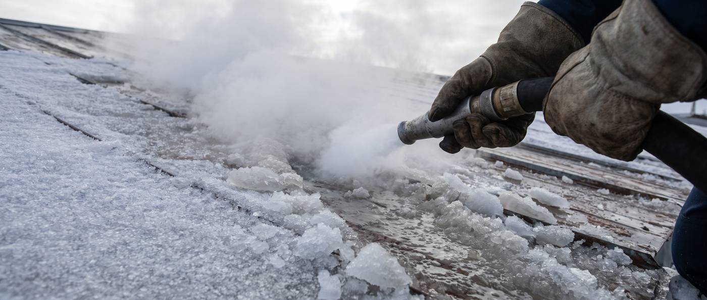 A low-pressure steam hose held by gloved hands melting a channel through thick roof ice